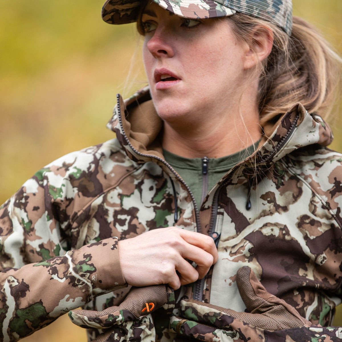 Woman zips up a hooded brown, green, and tan camouflage hunting jacket, wearing a matching cap and gloves with an orange logo.