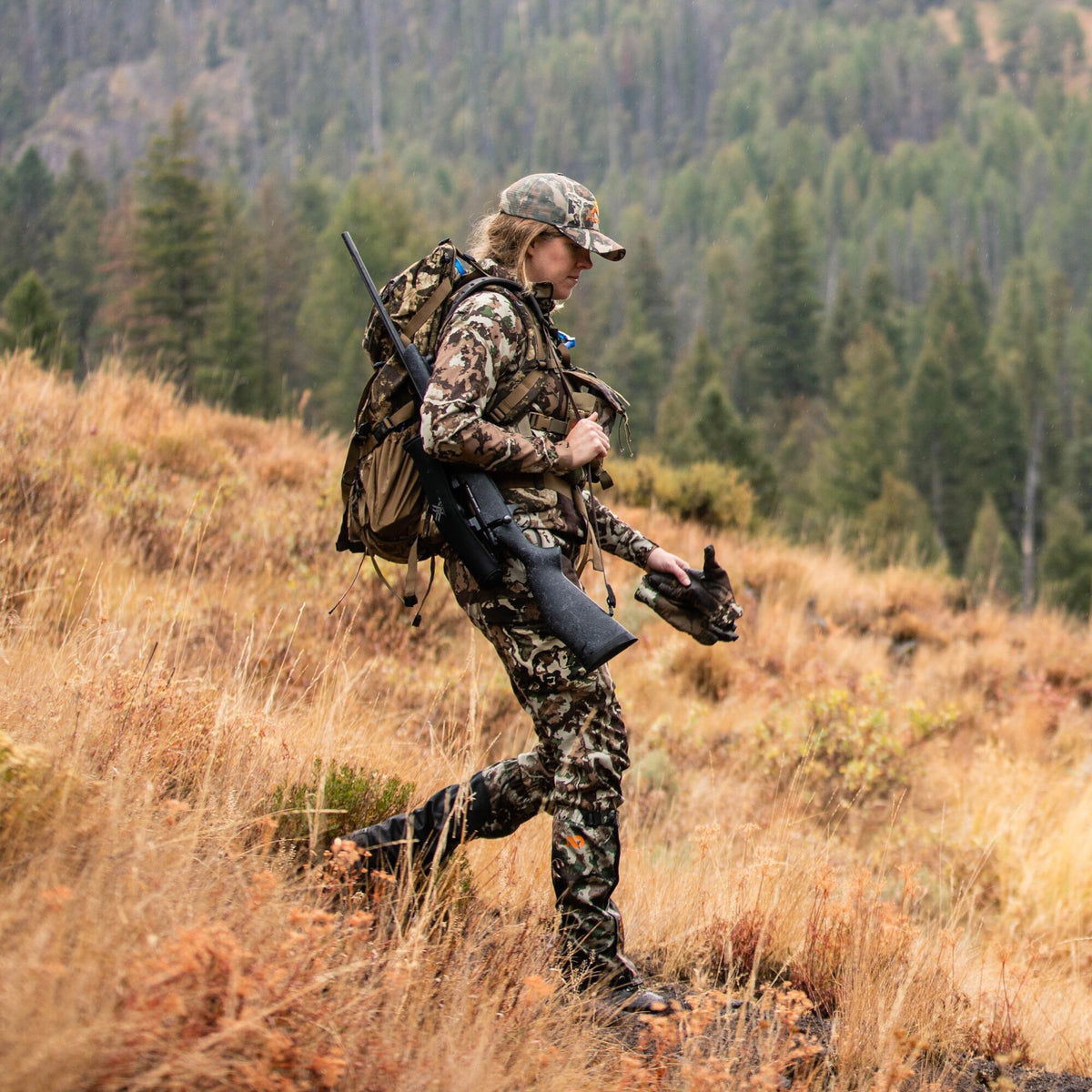 A woman in green and brown camouflage hunting gear, including a cap with an orange logo, carries a black rifle, tan backpack, and holds camouflage gloves while walking in a field.