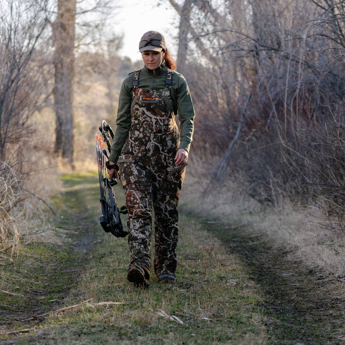 First Lite camouflage hunting bibs featuring chest straps and front pockets, worn by a woman holding a bow.