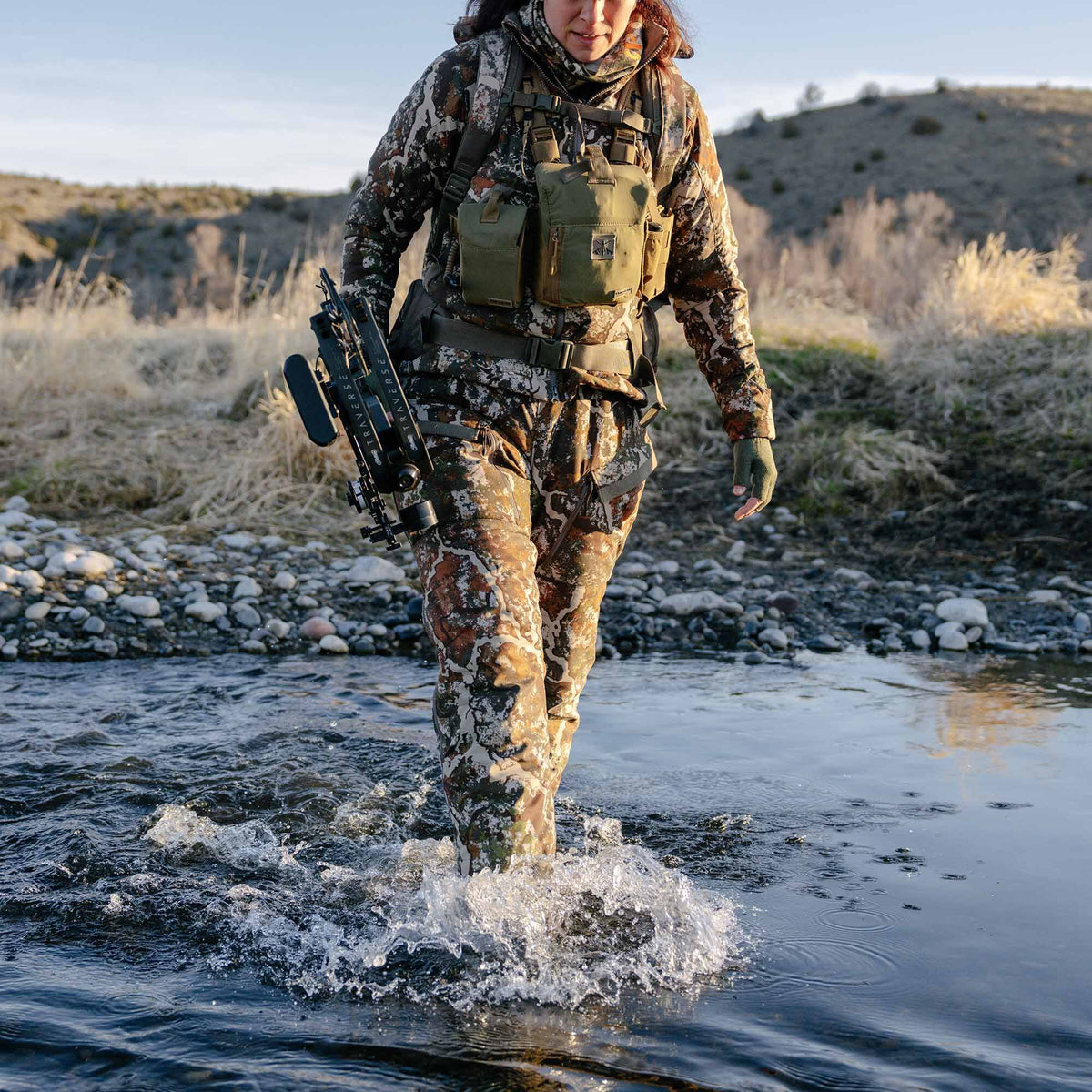 A person wears an earth-toned camouflage hunting suit with a chest rig and bow, wading through a rocky stream.