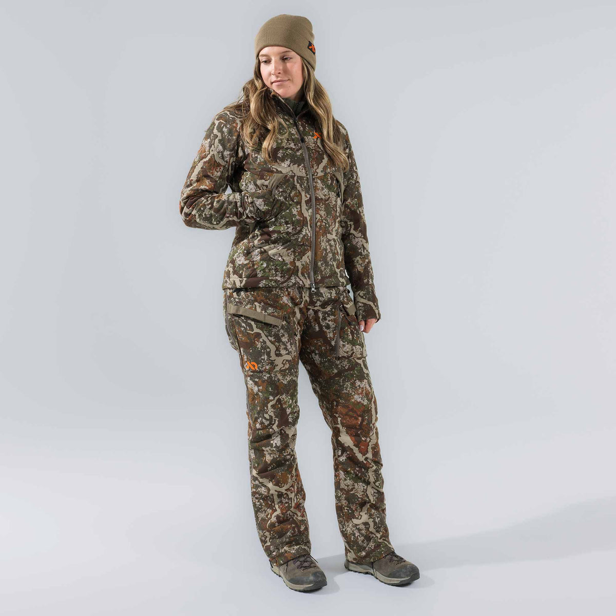 A woman models a full camouflage hunting suit, including a zippered jacket and cargo pants, an olive beanie, and hiking boots, with an orange logo visible.