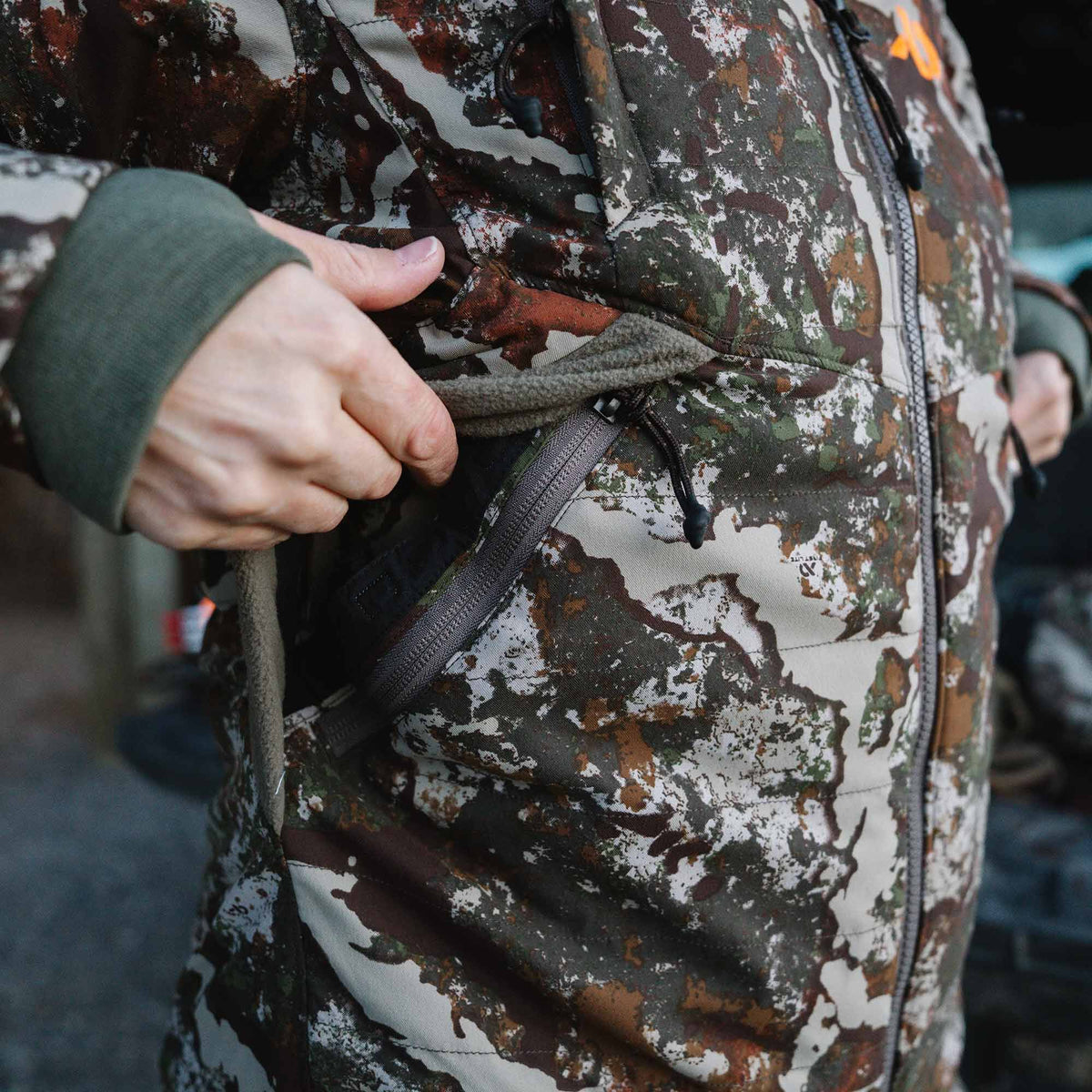 Close-up of a multi-terrain camouflage jacket with a hand opening a fleece-lined zippered pocket, showing a dark green cuff.