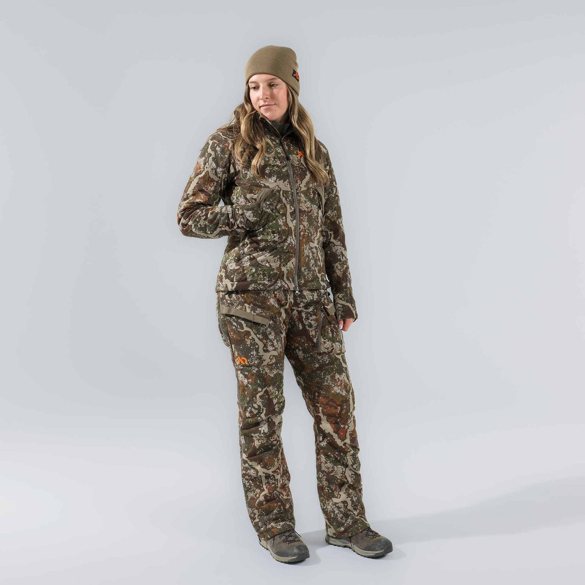 Women's camouflage hunting suit featuring a jacket and pants with orange logos, worn with an olive beanie and hiking boots.