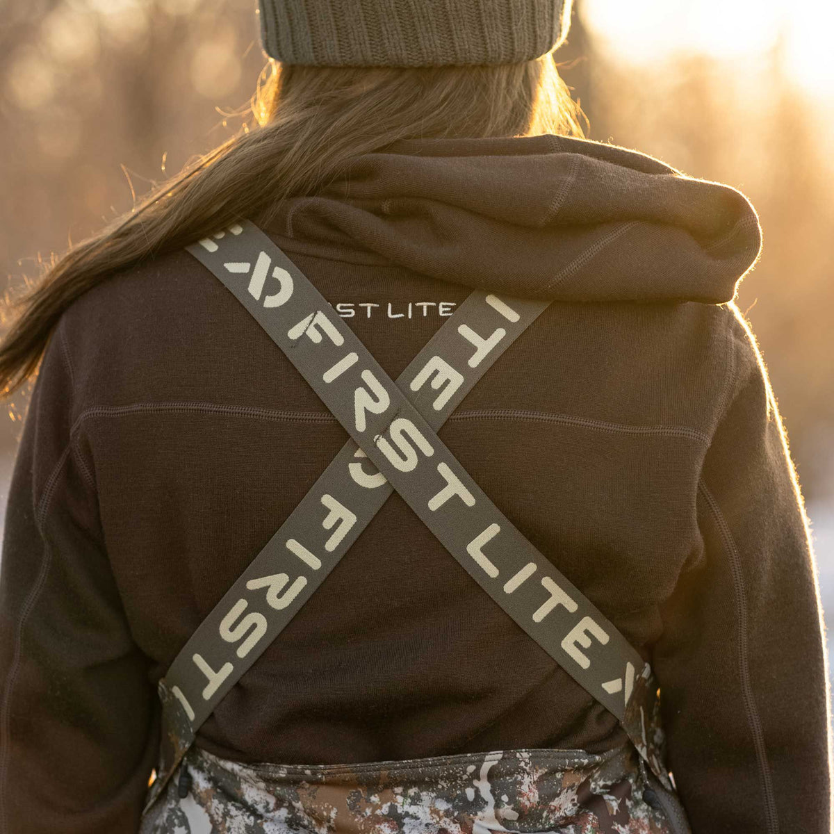 Back view of a person in a dark brown First Lite hooded sweatshirt with grey X-back suspenders over camouflage bibs, backlit by golden light.