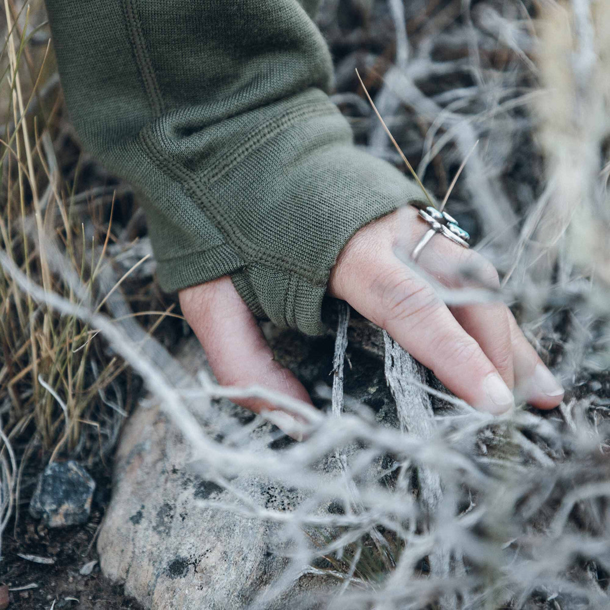 Olive green long-sleeve garment with a thumbhole cuff and prominent stitching, worn by a hand reaching into dry brush.