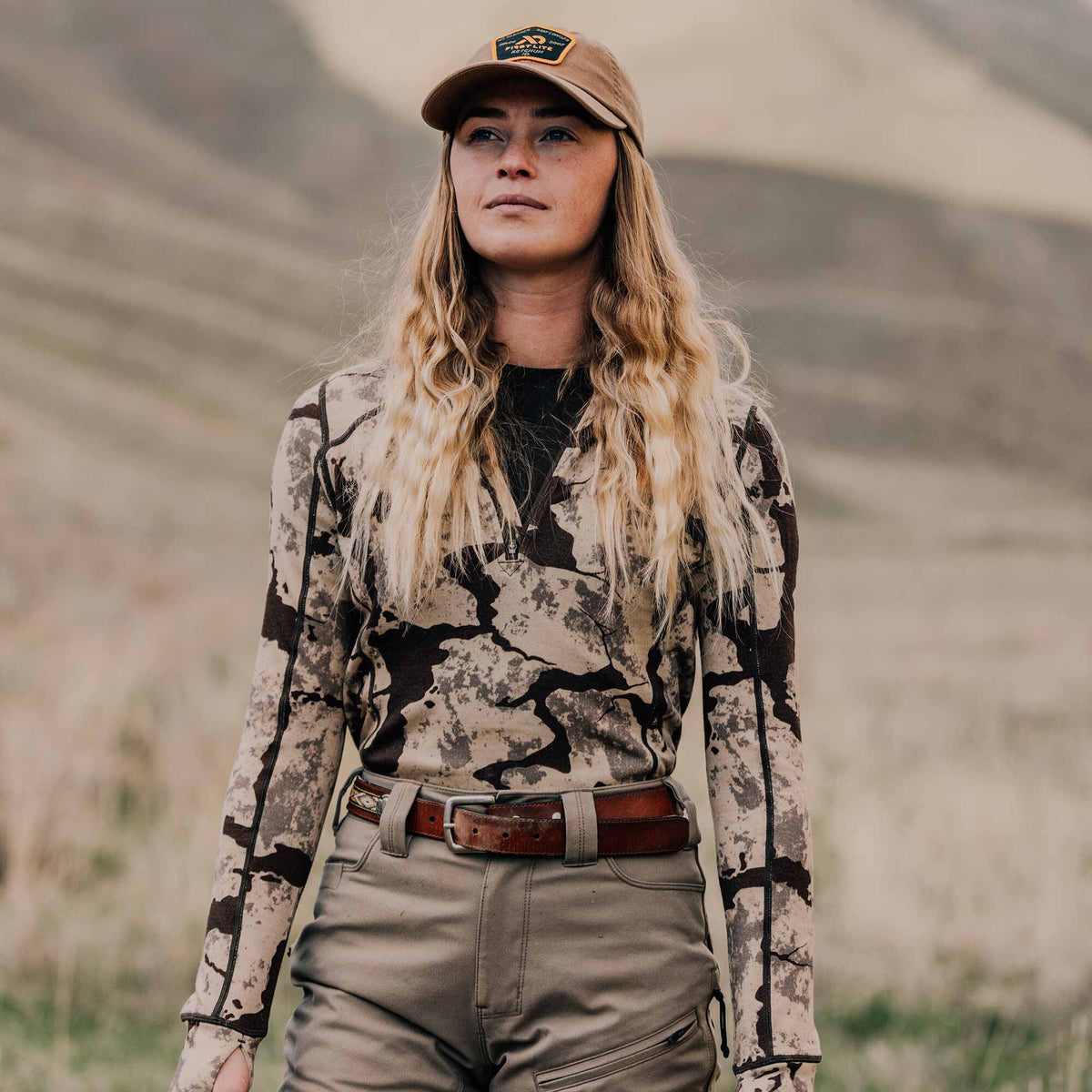 A woman models an earth-tone camouflage quarter-zip long-sleeve shirt with thumbholes, tan pants, a brown leather belt, and a tan cap.