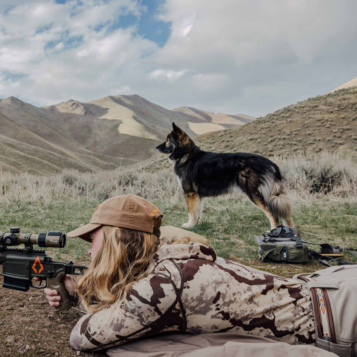 Dark grey and tan rifle with an orange logo and Sig Sauer scope, aimed by a person in a field.