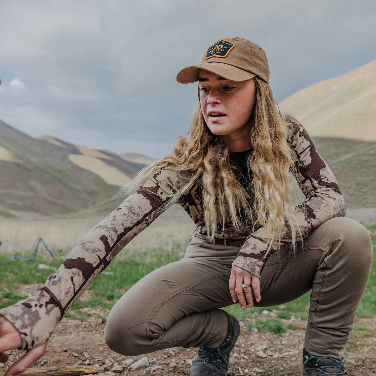 A woman crouches outdoors wearing a tan and brown camouflage hunting long-sleeve shirt with thumb loops, olive green pants, and a tan First Lite cap.