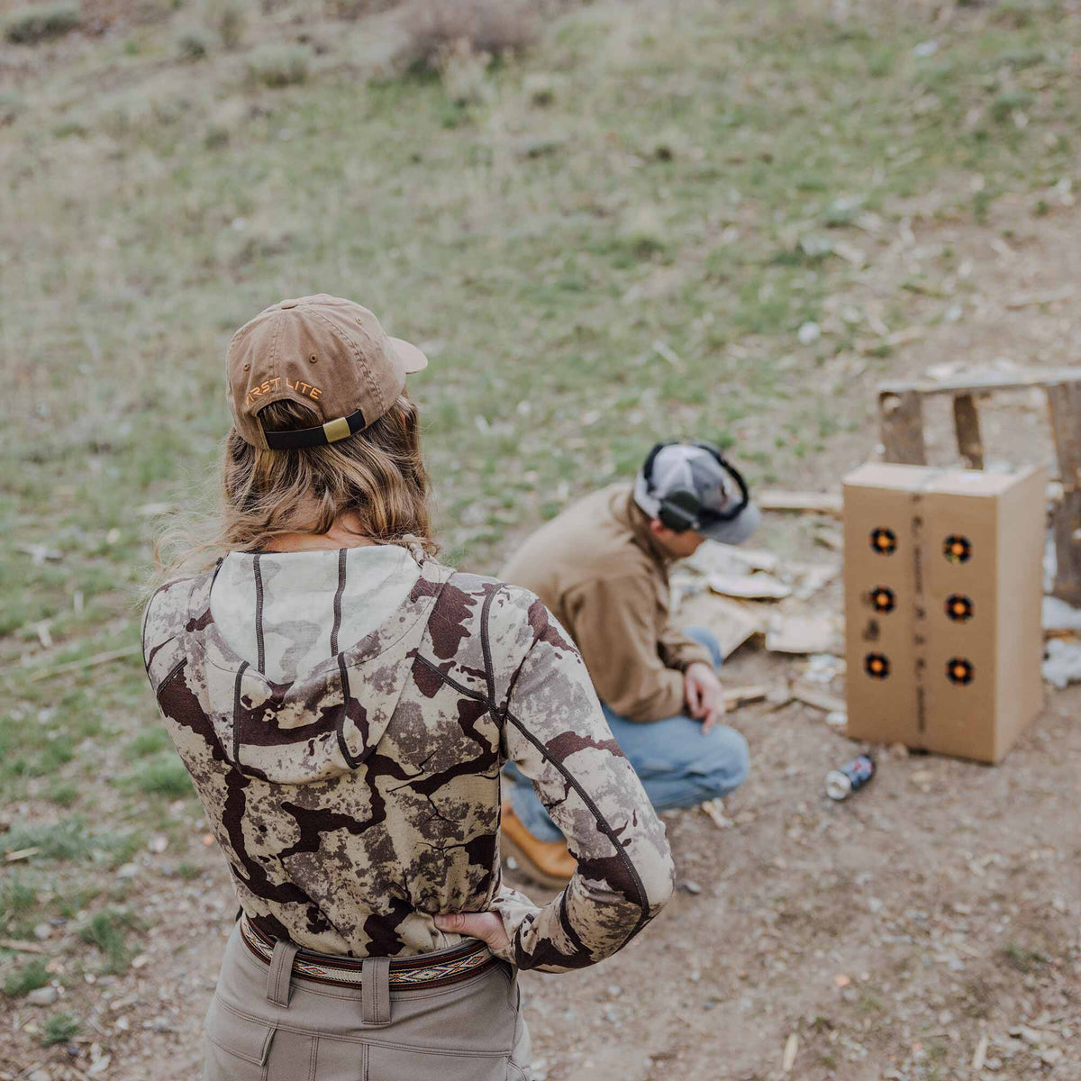 Brown and tan camouflage long-sleeve hooded shirt worn by a woman with a 'FIRST LITE' cap, standing outdoors.