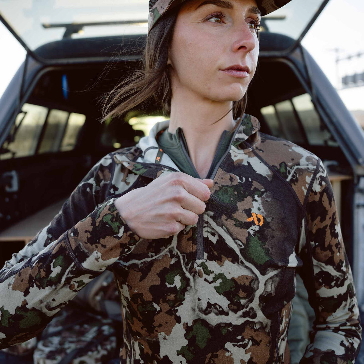 Woman wearing a camouflage 1/4-zip long-sleeved pullover with an orange logo on the left chest, zipping it up.