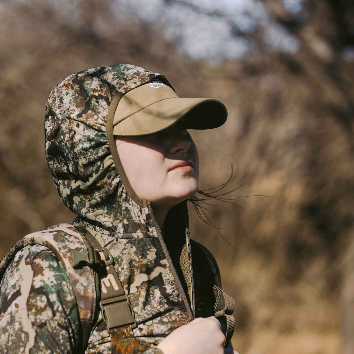 A person wears a multi-color camouflage hooded jacket, a tan baseball cap, and a chest harness with an "First Lite" logo.