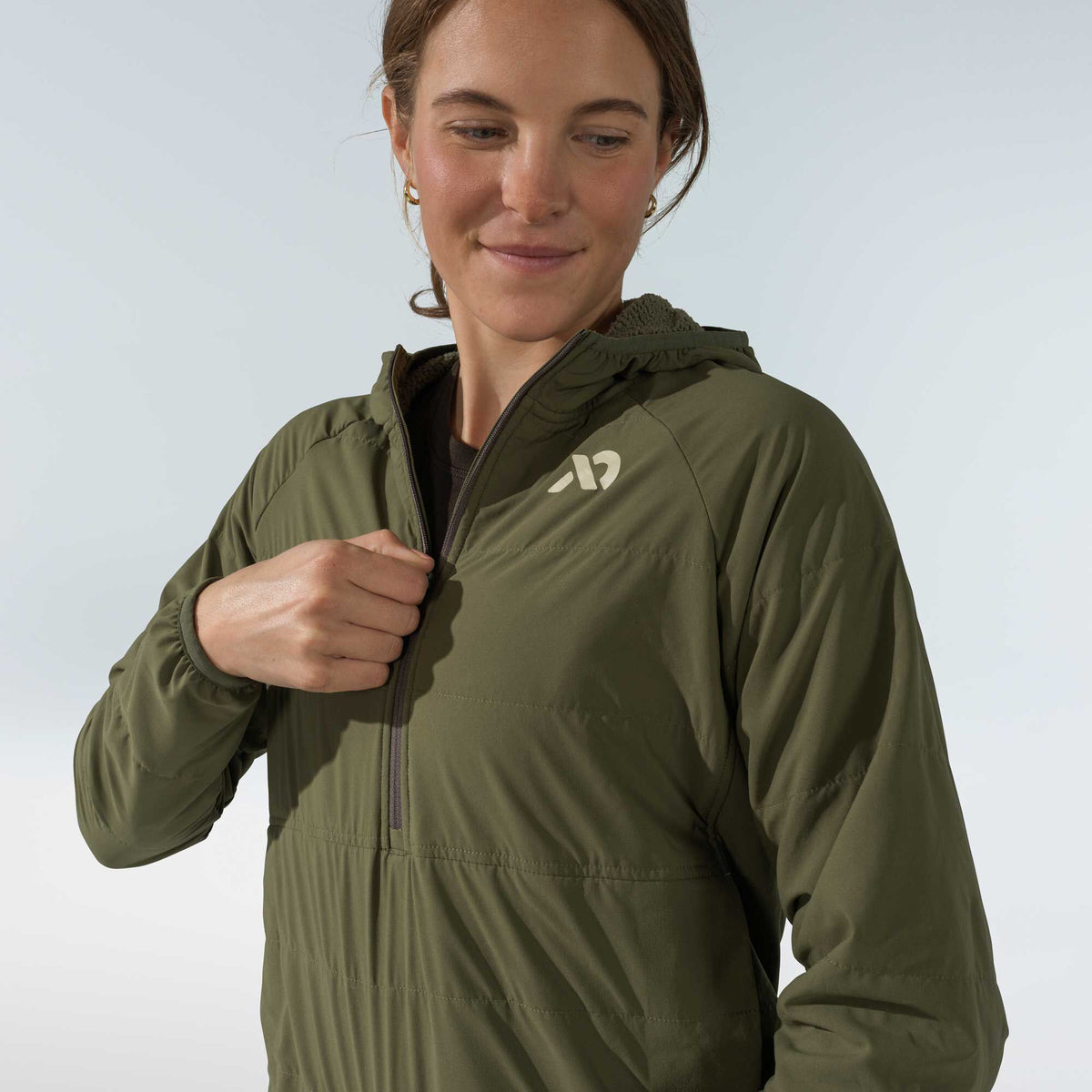 Olive green hooded quarter-zip jacket with a chest logo, worn by a woman zipping it up.