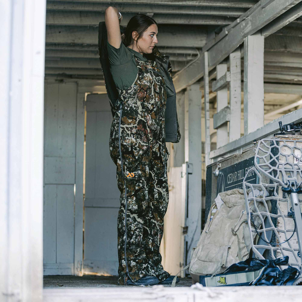 Woman in mottled brown and green camouflage hunting bibs with full-length leg zippers, putting on a jacket.