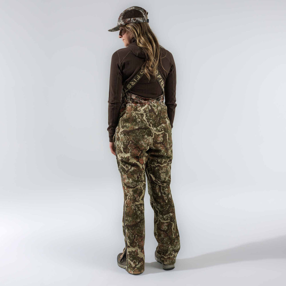Camouflage hunting bibs with 'FIRST LITE' logo straps and full-length side leg zippers, modeled from behind.