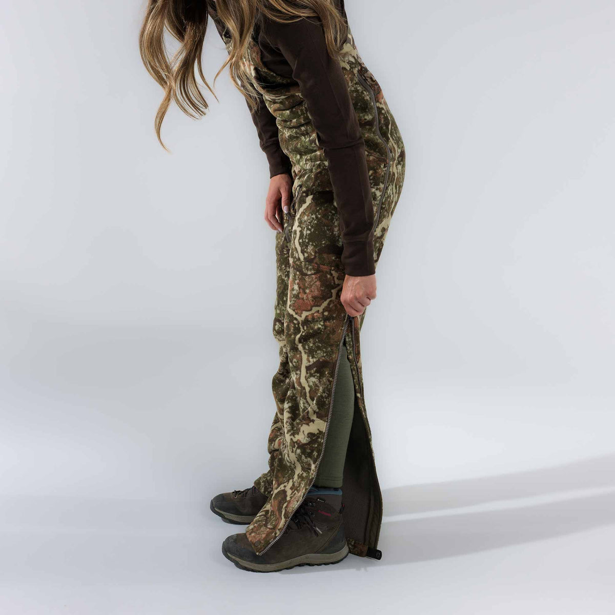 Camouflage hunting bib overalls with full-length side zippers, one partially unzipped revealing a green inner layer.