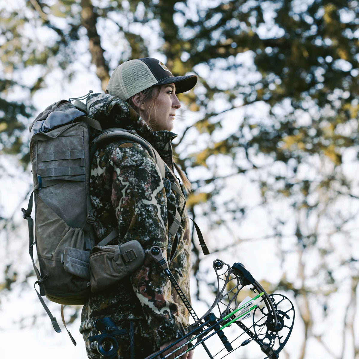 A woman in camouflage jacket and mesh hat, wearing a dark grey hunting backpack with MOLLE webbing, and holding a black compound bow with green arrows.