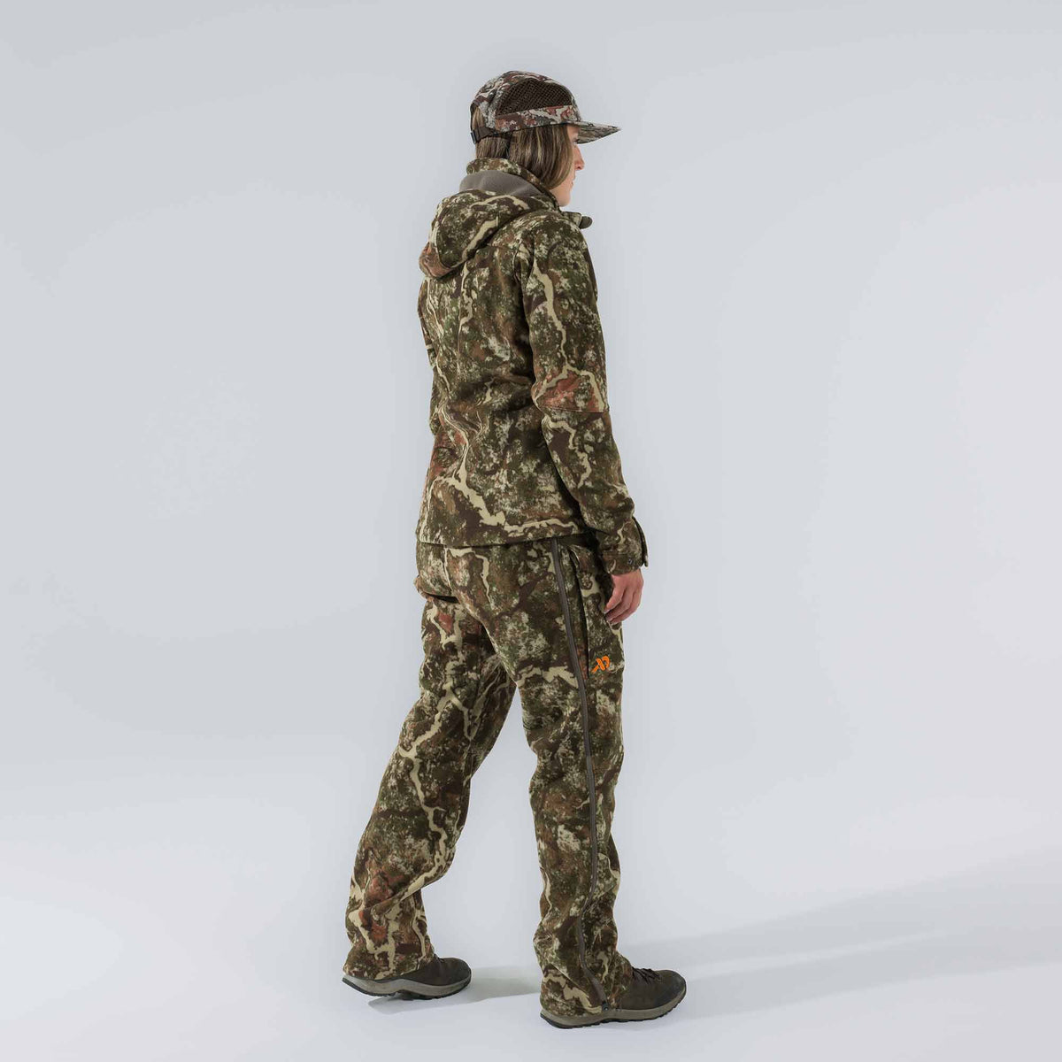 A person wears a full green, brown, and tan camouflage hunting outfit with a hooded jacket, pants featuring full-length side zippers, and a mesh-back cap, showing an orange logo on the right thigh.
