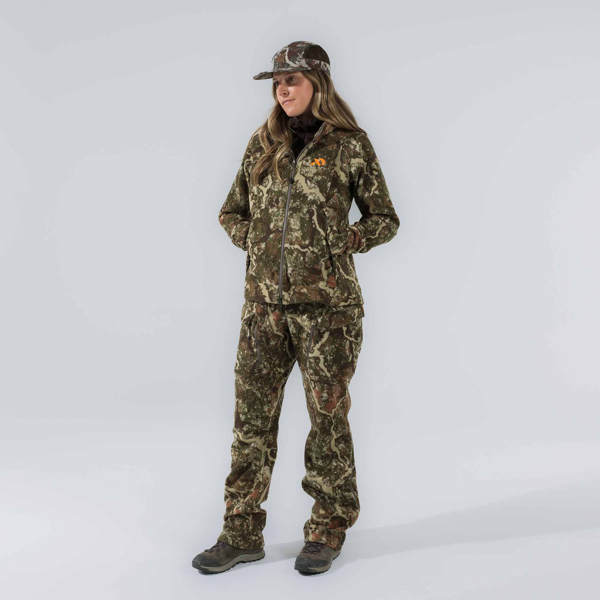 Woman wearing a full green and brown camouflage hunting outfit, including a zippered jacket with an orange logo, pants with zippered pockets, and a mesh-back cap.