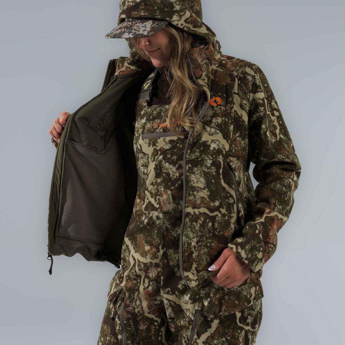 Woman models a multi-colored camouflage fleece hunting suit, featuring a hooded jacket open to reveal its dark green mesh lining, and bib overalls with an orange "First Lite" logo.