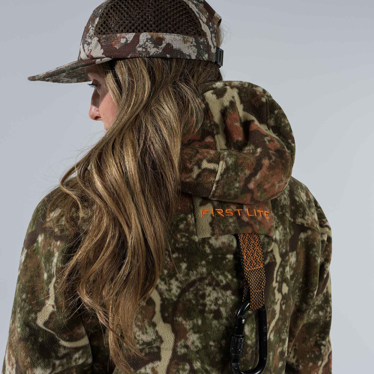 A woman wears a First Lite camouflage fleece hoodie featuring an orange logo and attached carabiner, along with a matching mesh-back cap.