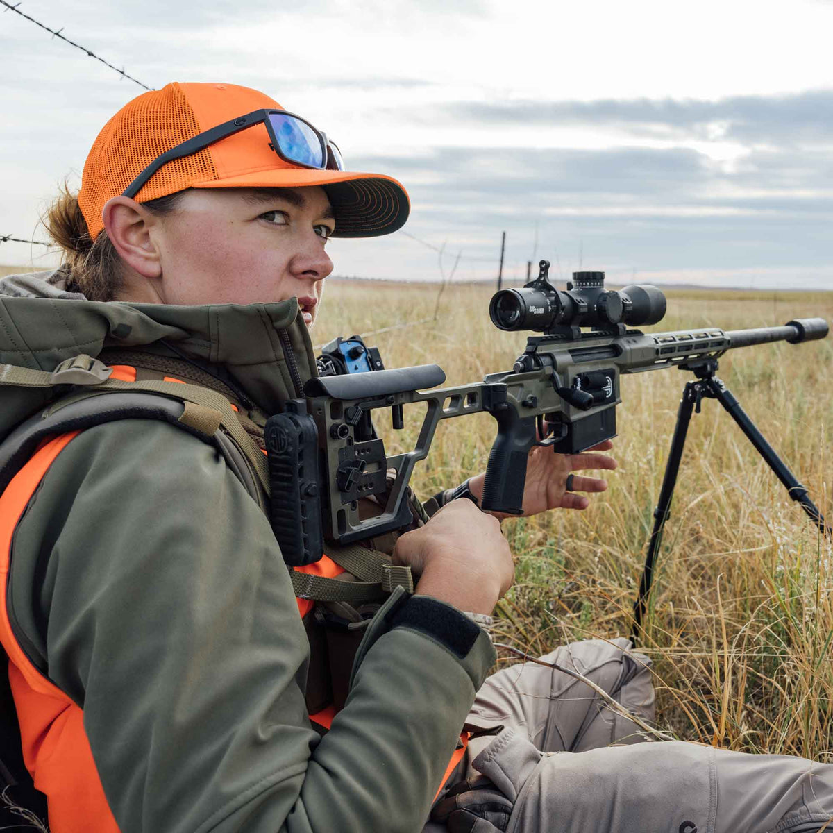 Olive green tactical rifle with a Sig Sauer scope, suppressor, and adjustable skeletonized stock, supported on a tripod.
