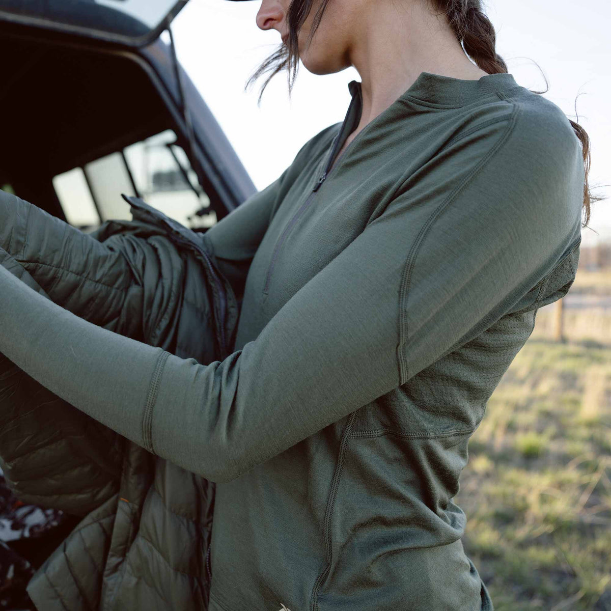A woman wears an olive green quarter-zip long-sleeve top featuring a mock neck and visible flatlock seams.