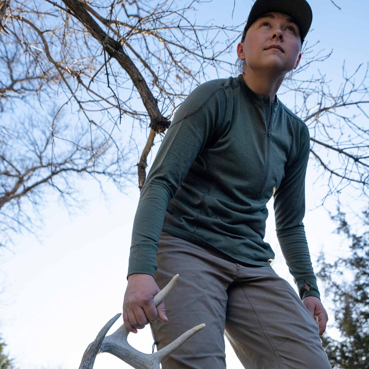 A woman wears an olive green quarter-zip long-sleeve shirt with thumbholes and tan pants, holding an antler outdoors.