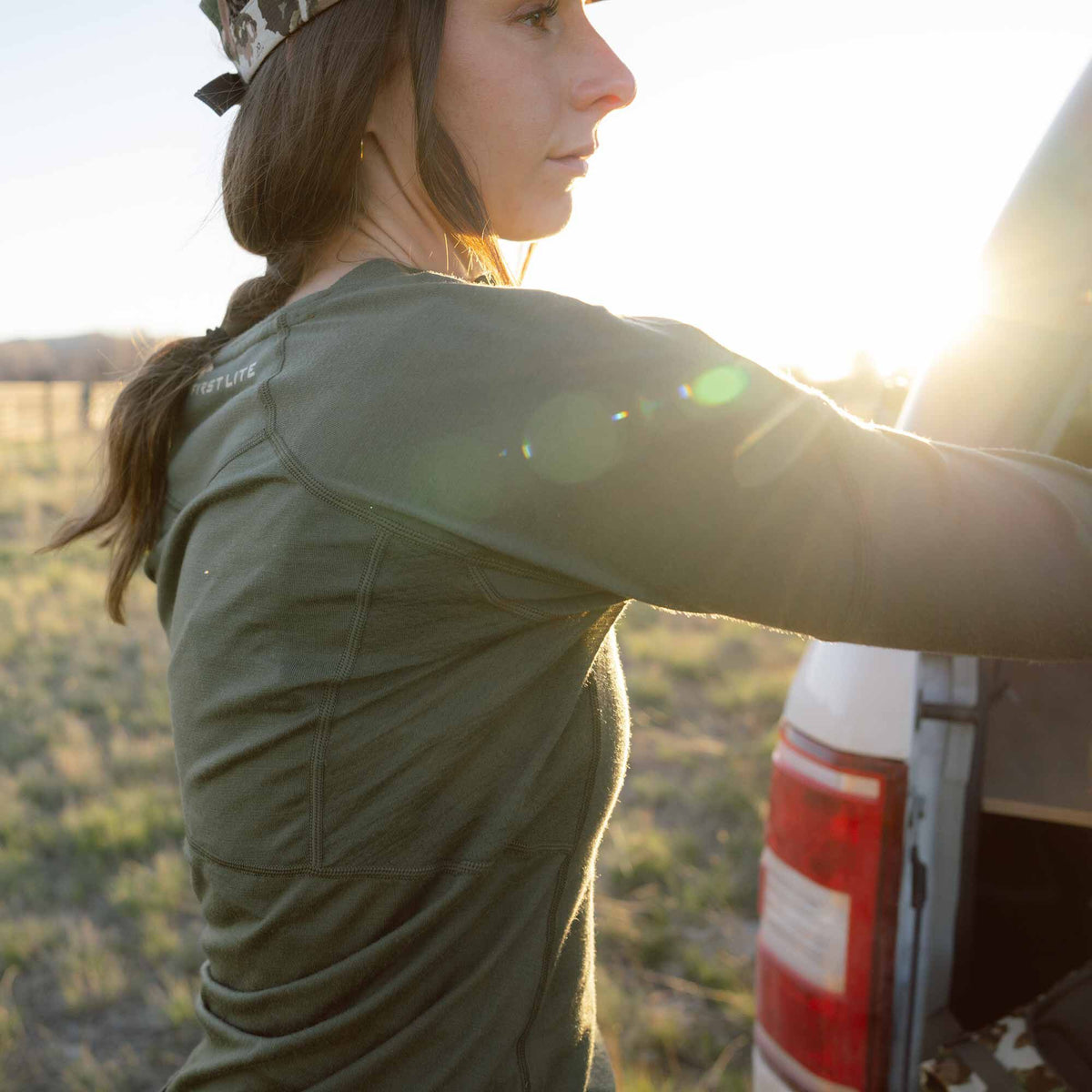 Woman wearing a form-fitting olive green long-sleeve base layer with a "FIRST LITE" logo visible on the shoulder.
