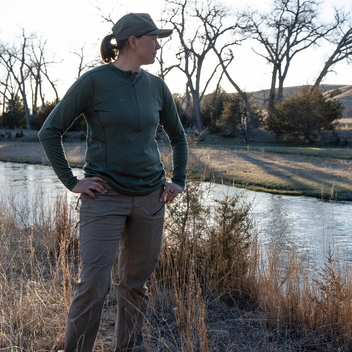 A woman wears a dark olive green quarter-zip long-sleeve top, matching baseball cap, and tan outdoor pants by a river.
