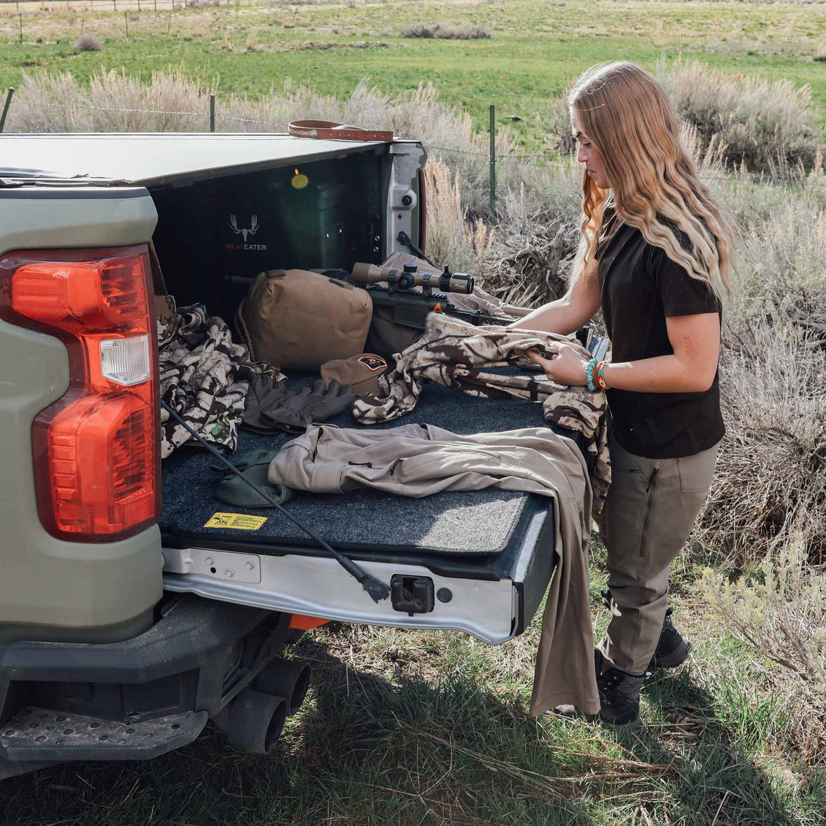 A woman organizes hunting gear on a dark gray carpeted truck bed slide-out system, extended over the open tailgate of a light green pickup.