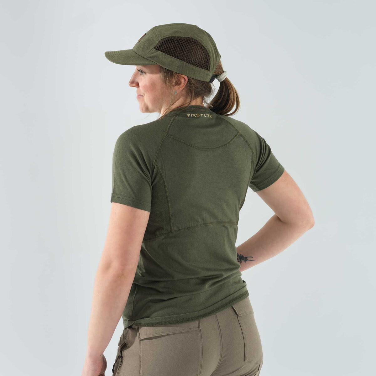 A woman wears an olive green First Lite short-sleeved t-shirt with raglan sleeves and a matching cap featuring mesh side panels.