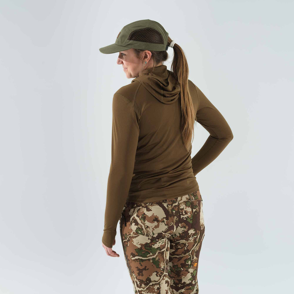 A woman wears an olive green mesh cap, an olive brown hooded long-sleeve shirt, and earth-toned camouflage pants.