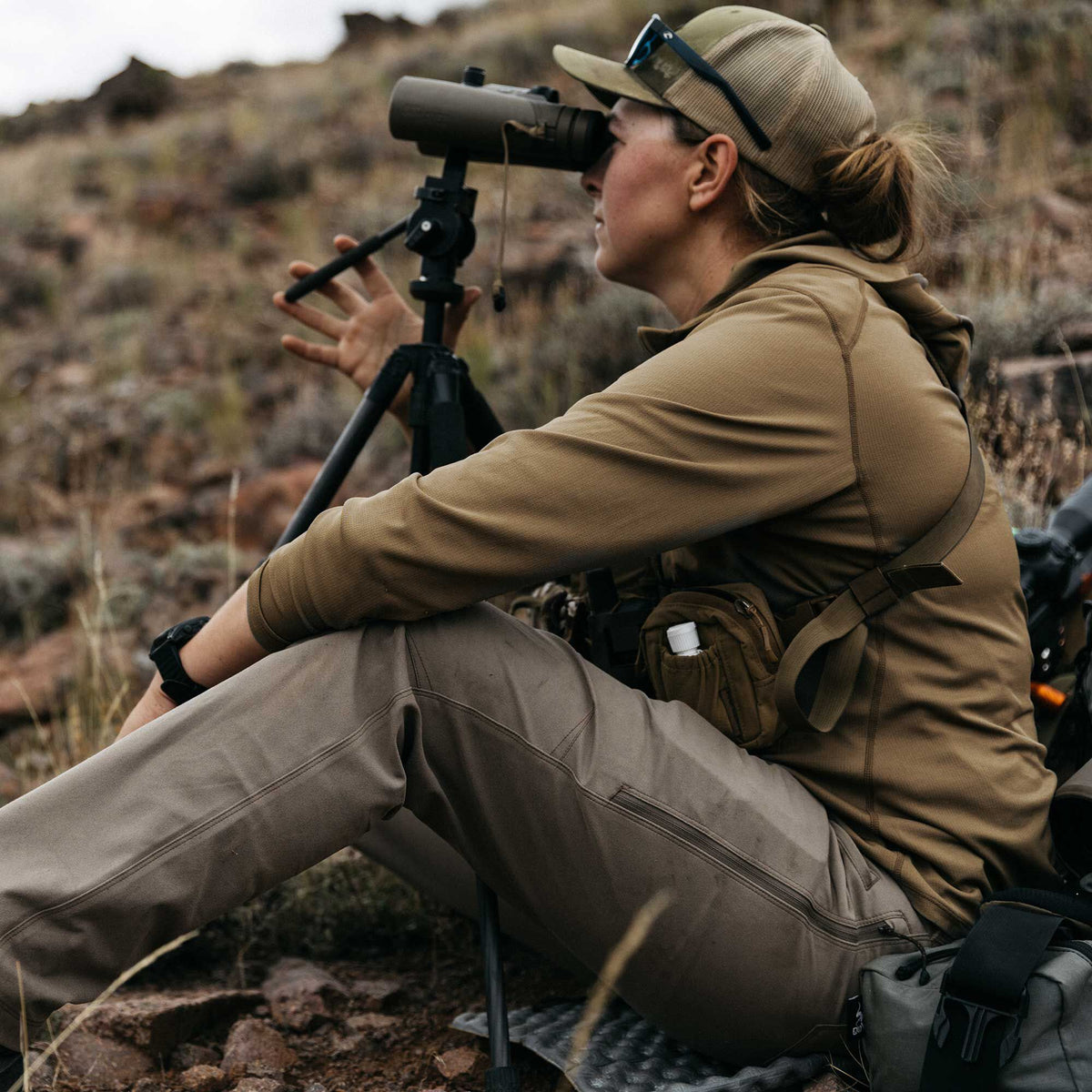 A woman wears a tan long-sleeved performance shirt with flatlock seams and khaki outdoor pants with a zippered thigh pocket.
