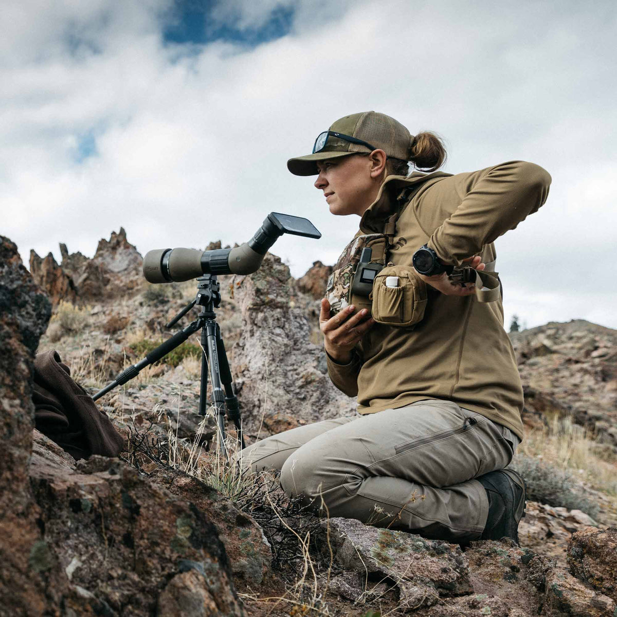 A woman in a tan shirt and olive cap kneels on rocky ground, wearing a tan chest rig with binoculars, beside a dark green spotting scope with a smartphone attached to a black tripod.