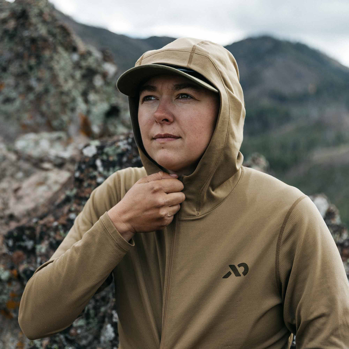 A woman wears a tan waffle-knit long-sleeve hoodie with a black chest logo and a cap under the hood, adjusting the partial zipper.