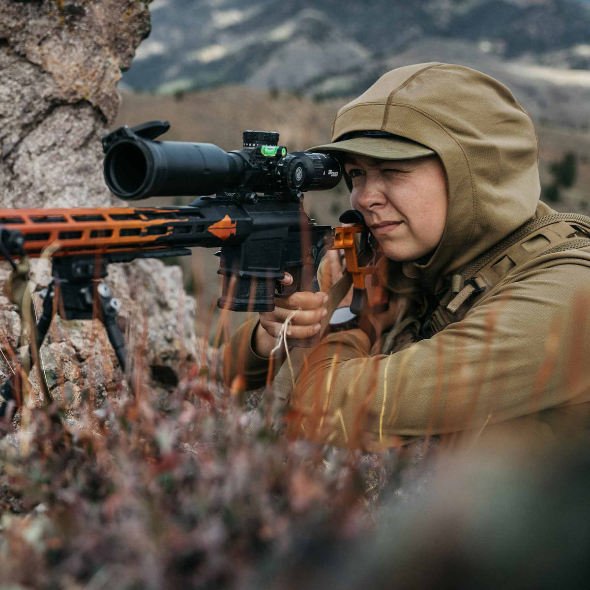 Orange and black precision rifle with a scope, green level, bipod, and orange support bag, aimed by a shooter.
