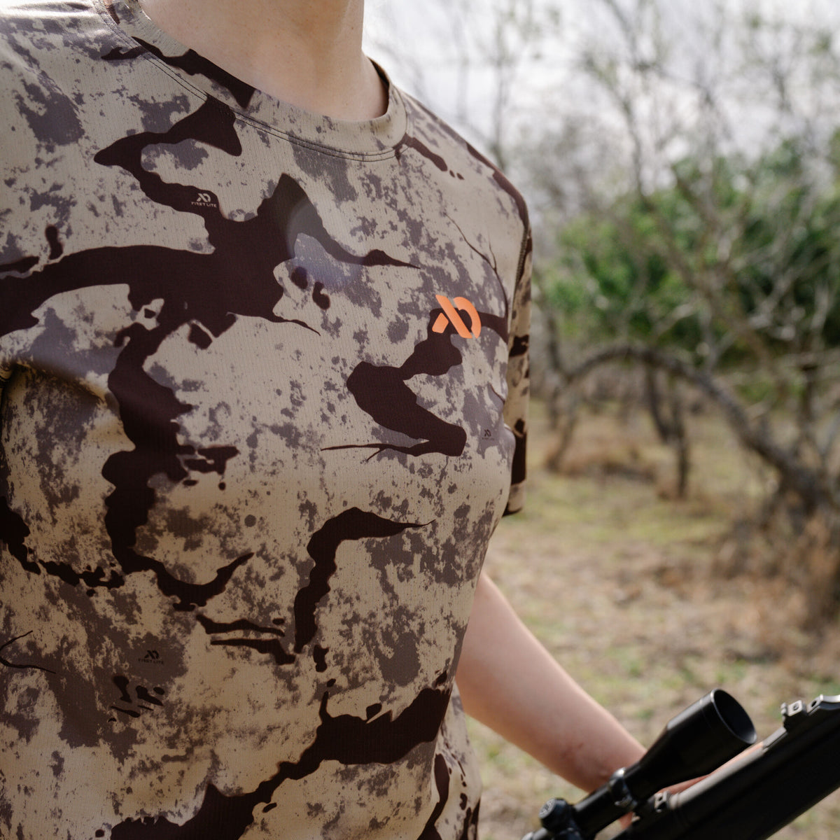 A camouflage t-shirt with beige and dark brown patterns, featuring a prominent orange 'X' logo on the chest and a smaller First Lite logo on the upper left, worn by a person holding a rifle outdoors.