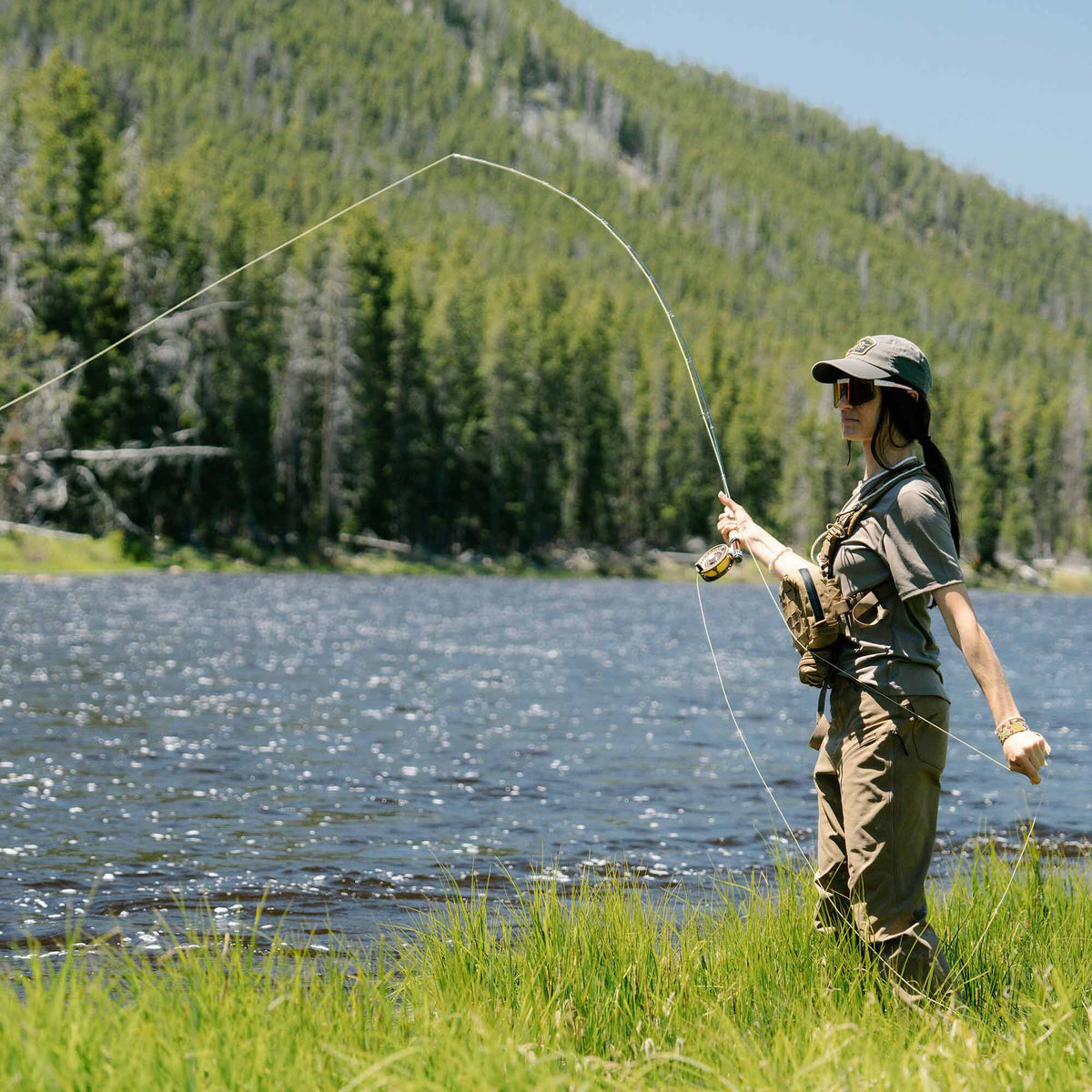 Woman in a khaki fishing outfit and hat casts a fly rod with a yellow reel on a lake.