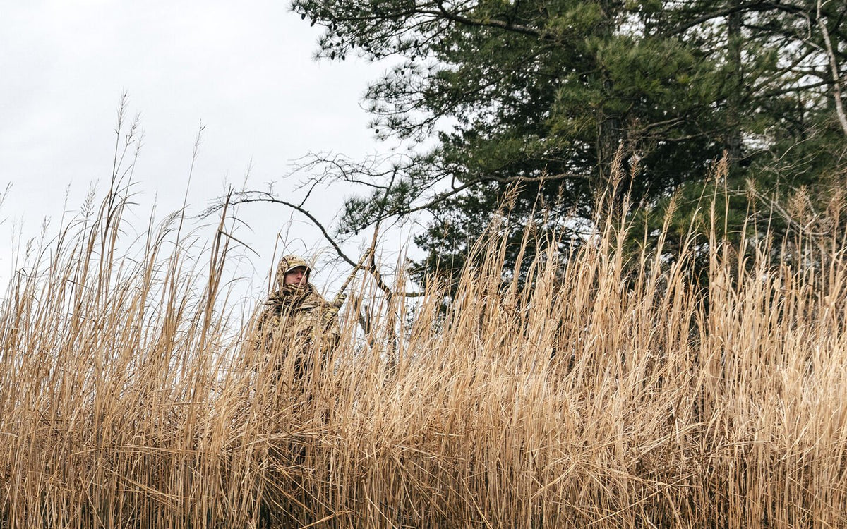 Tan camo gear on person, holding rifle, blending into dry tall grass.